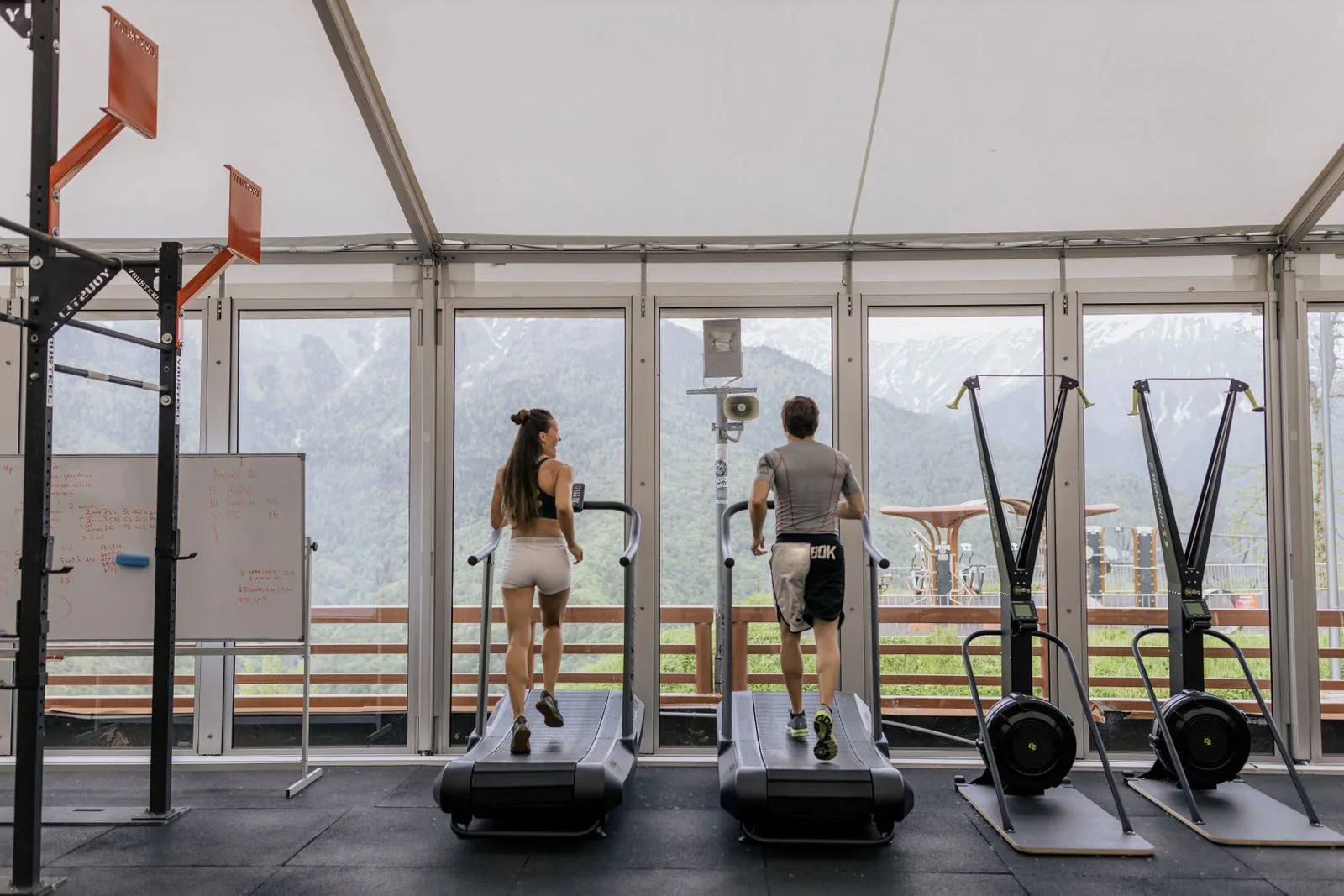 Zwei Personen auf Laufbändern in einem Fitnessstudio mit Bergblick durch die Fenster